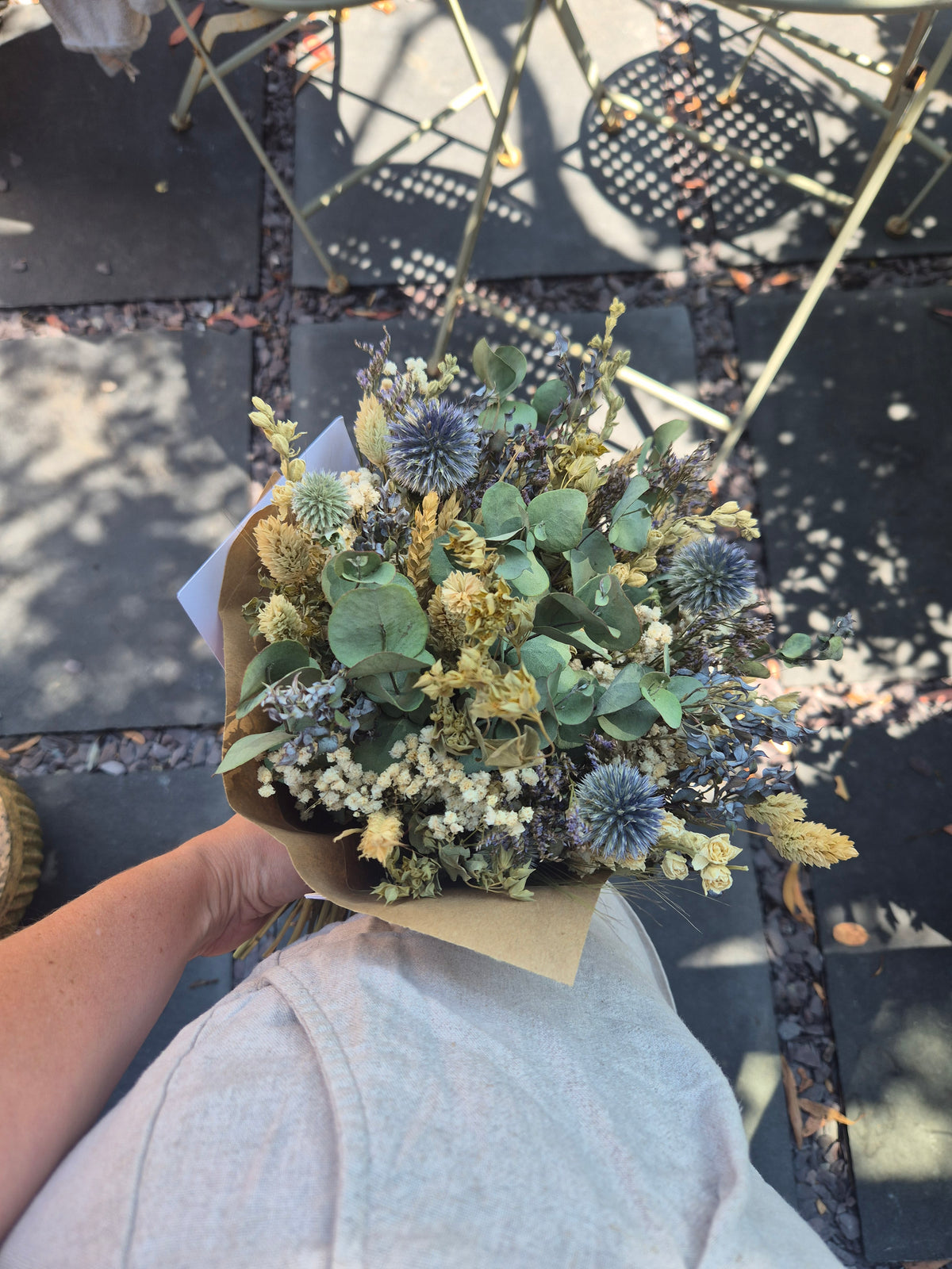 A dried flower bouquet wrapped in brown kraft paper and held by a person wearing a light gray t-shirt, photographed from a top-down perspective on a dark stone patio.