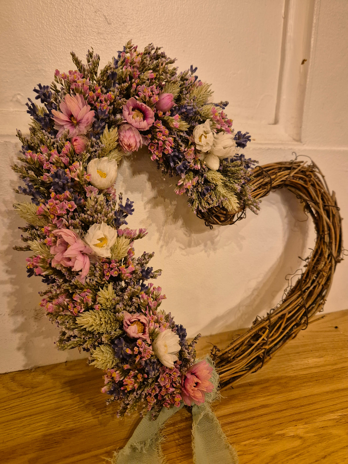 Dried flower wreath in the shape of a heart with half decorated in pink, blue and green flowers and foliage