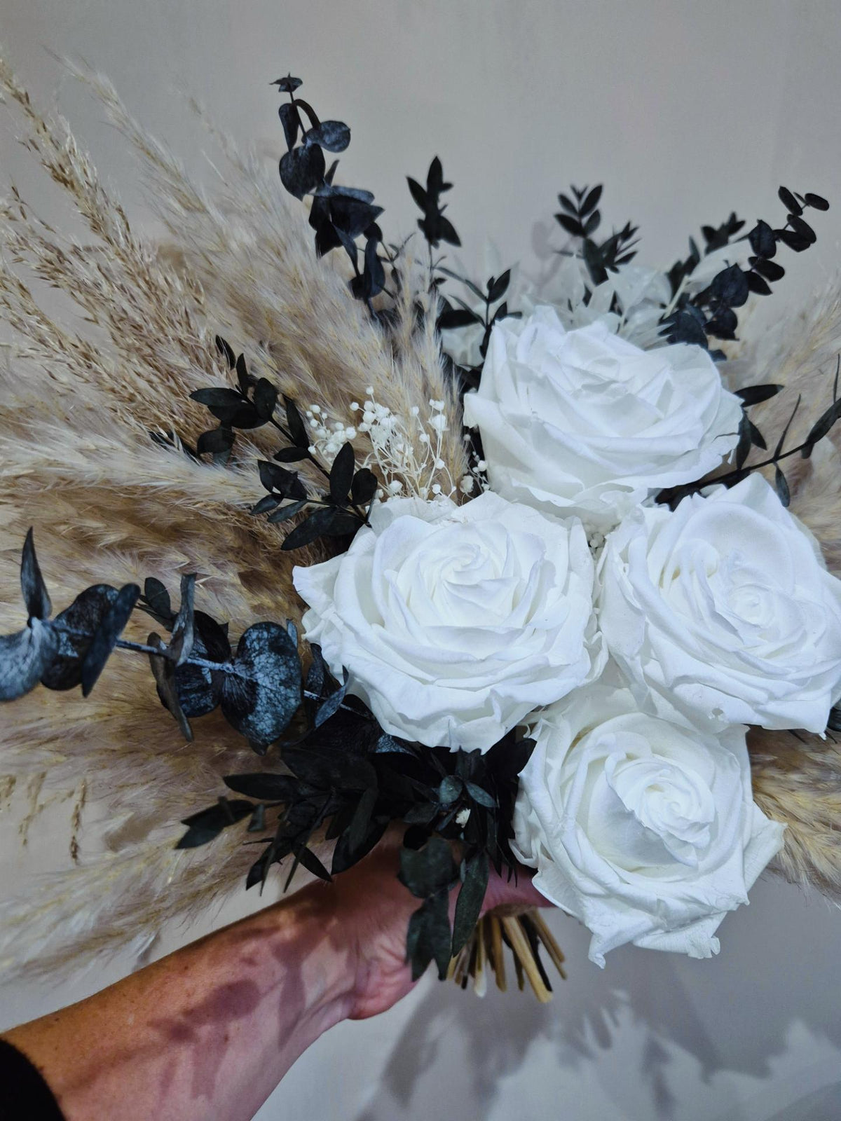 A brides dried floral bouquet featuring four large white roses, beige pampas grass, and dark green to black eucalyptus leaves. A boho inspired bouquet.