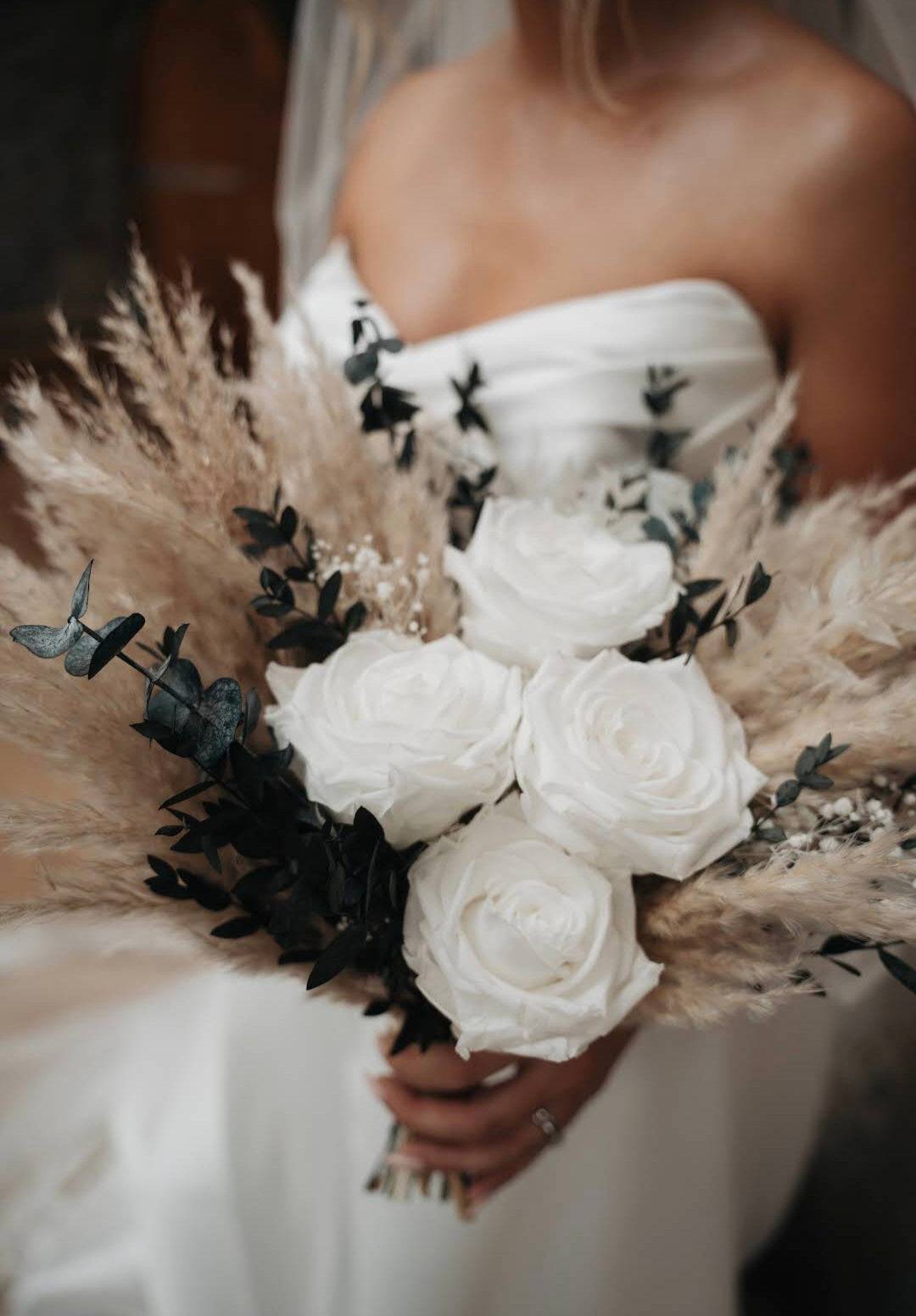 A close-up of a bride holding a bridal bouquet featuring white roses, beige pampas grass, and dark green eucalyptus leaves.