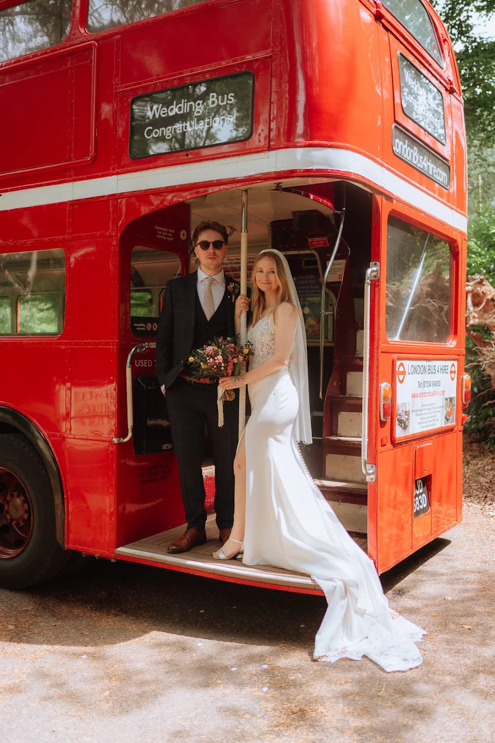 A bride in a white wedding gown and a groom in a dark suit stand on the open back of a red double-decker bus, holding a bouquet. The bus has a custom 'Wedding Bus Congratulations' sign on the front destination panel.