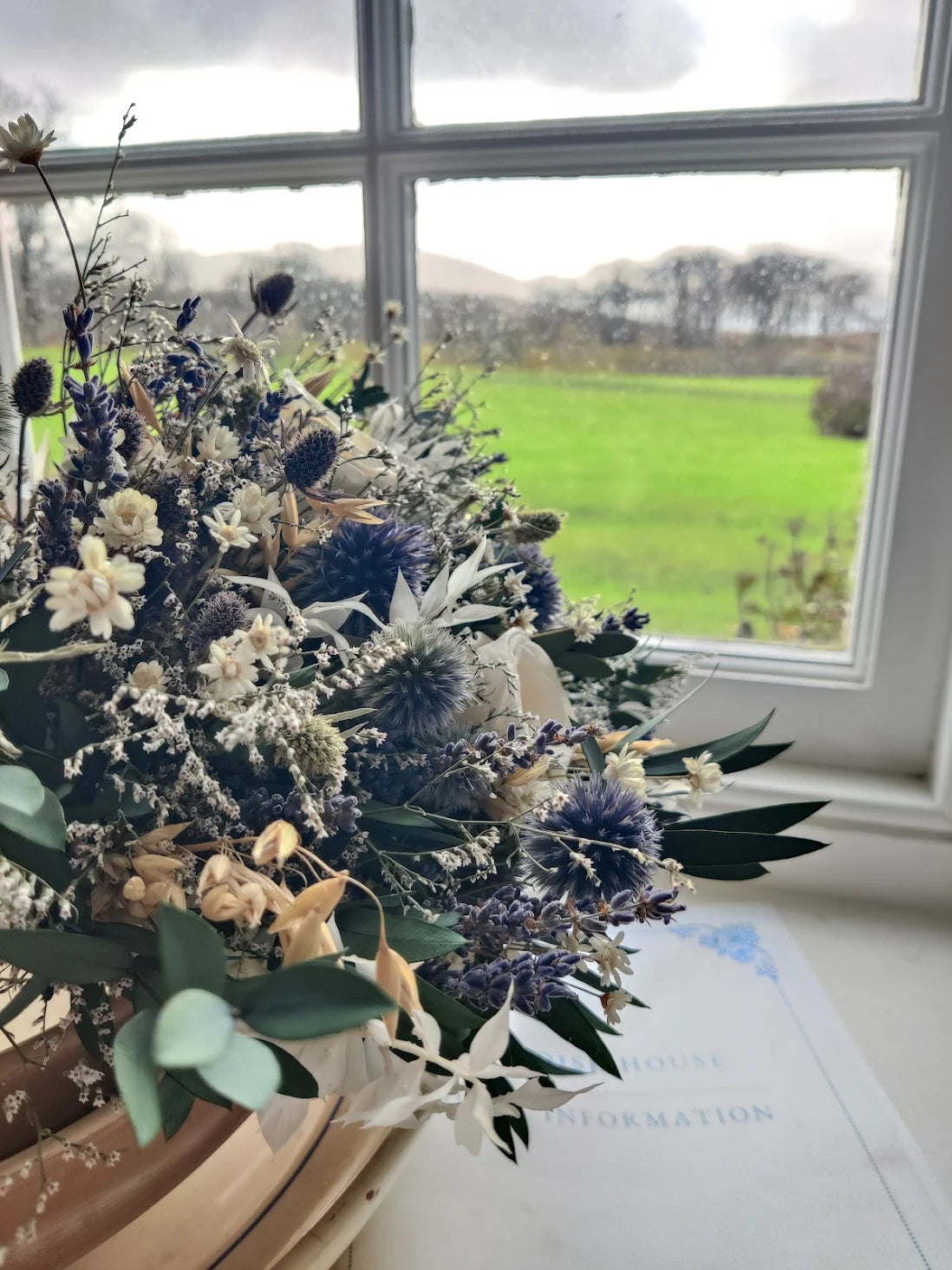 A dried flower arrangement featuring blue thistles, white flowers, and green foliage, displayed in a wooden pot on a windowsill with a scenic green field visible outside.