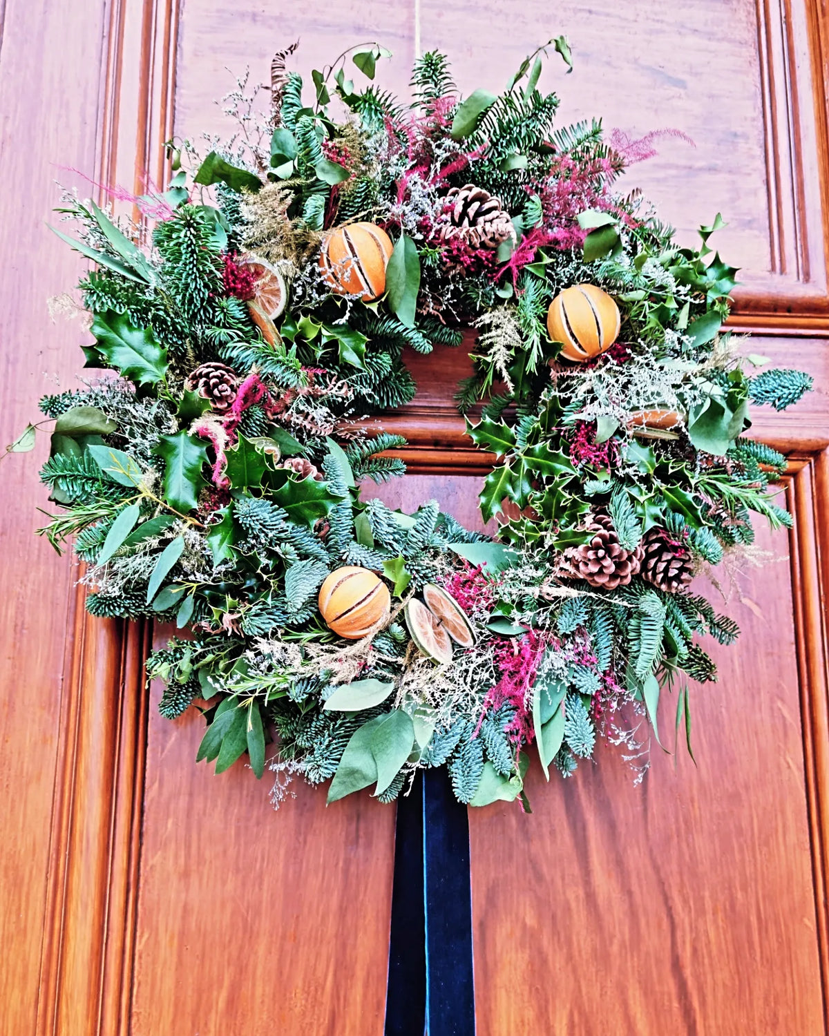 A festive holiday wreath hanging on a warm brown wooden door. The wreath is made of green evergreen foliage, pinecones, dried orange slices, and small orange basketball ornaments.