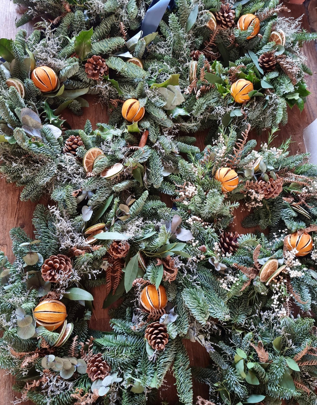A close-up of a rustic Christmas wreath made of green pine and fir branches, decorated with small whole oranges, pinecones, dried lime slices, and eucalyptus leaves on a wooden surface.