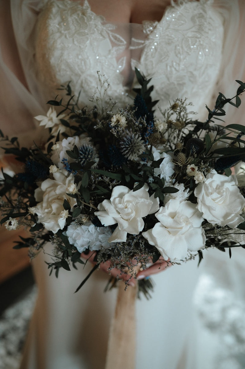 A close up image of a brides bouquet of stunning preserved gardenia roses, hydrangea and eucalyptus against a sparkling dress. 
