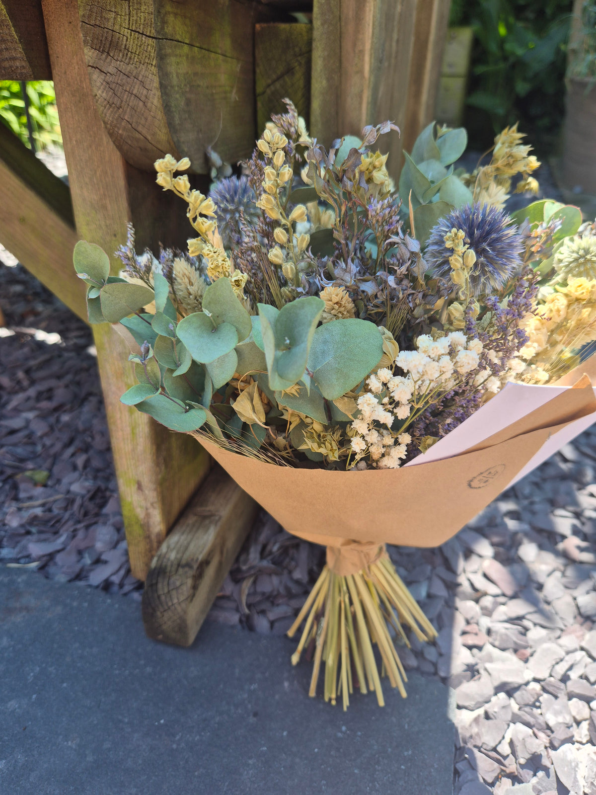 A dried flower bouquet wrapped in brown kraft paper, featuring green eucalyptus leaves, purple thistle-like blooms, and gypsophila, resting on a wooden bench outdoors.