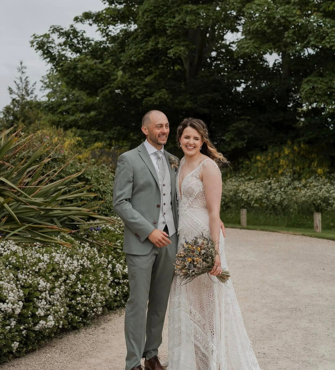 A bride in a sleeveless lace wedding gown holding a bouquet of wildflowers stands next to a groom in a light gray suit on a gravel path surrounded by lush green trees and white flowers.