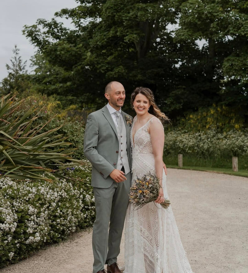 A bride in a sleeveless lace wedding gown holding a bouquet of wildflowers stands next to a groom in a light gray suit on a gravel path surrounded by lush green trees and white flowers.