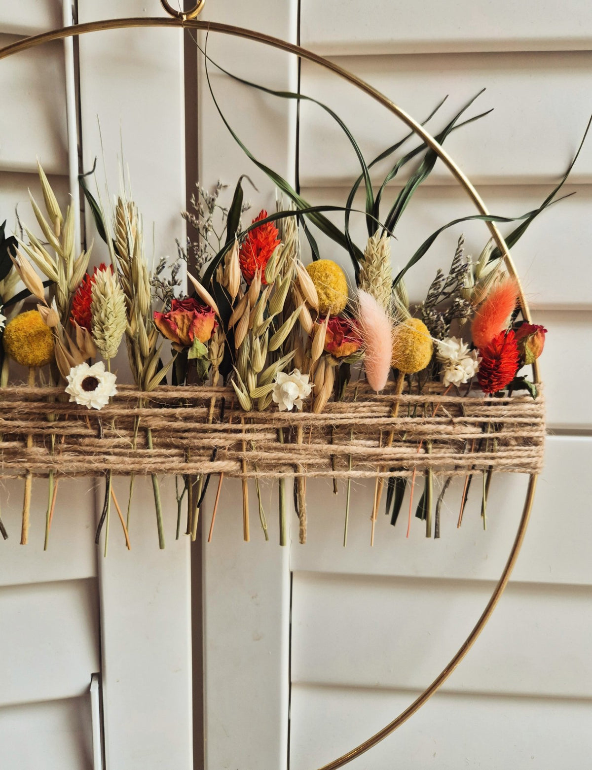 A rustic dried flower and grass arrangement in a woven hoop with twine, hanging inside a large gold-toned circular frame. The arrangement includes red, orange, yellow, beige, and green botanical elements.
