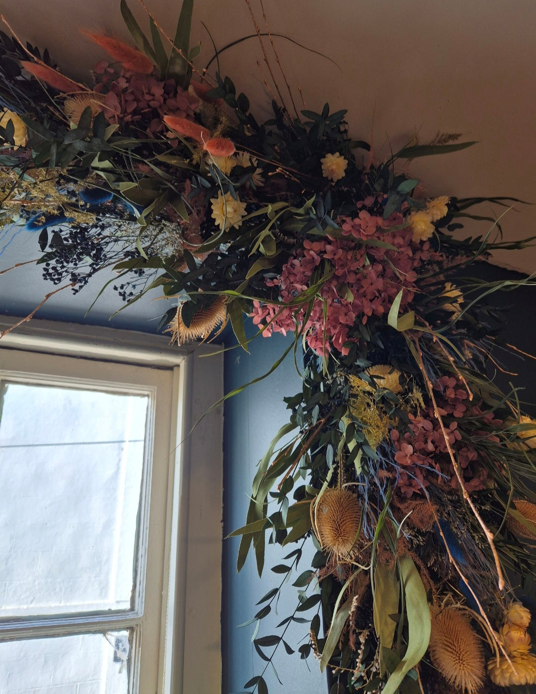 A large, rustic dried flower and botanical garland hanging in the corner of a room, featuring pink flowers, green foliage, brown seed pods, and yellow accents.