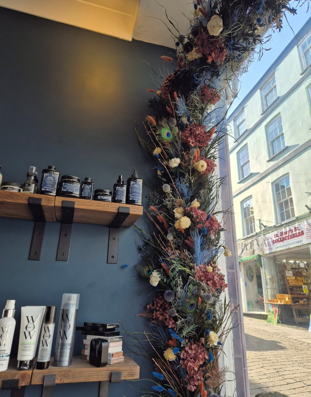 A tall decorative floral garland with dried flowers, peacock feathers, and botanical stems hangs vertically beside a window in a salon or retail interior. 