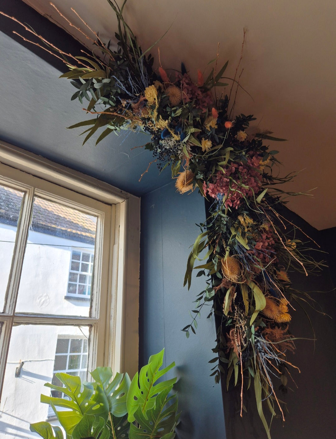 A large, rustic dried flower window display with green foliage, thistles, and hydrangeas, draped along the ceiling and corner of a room next to a window.