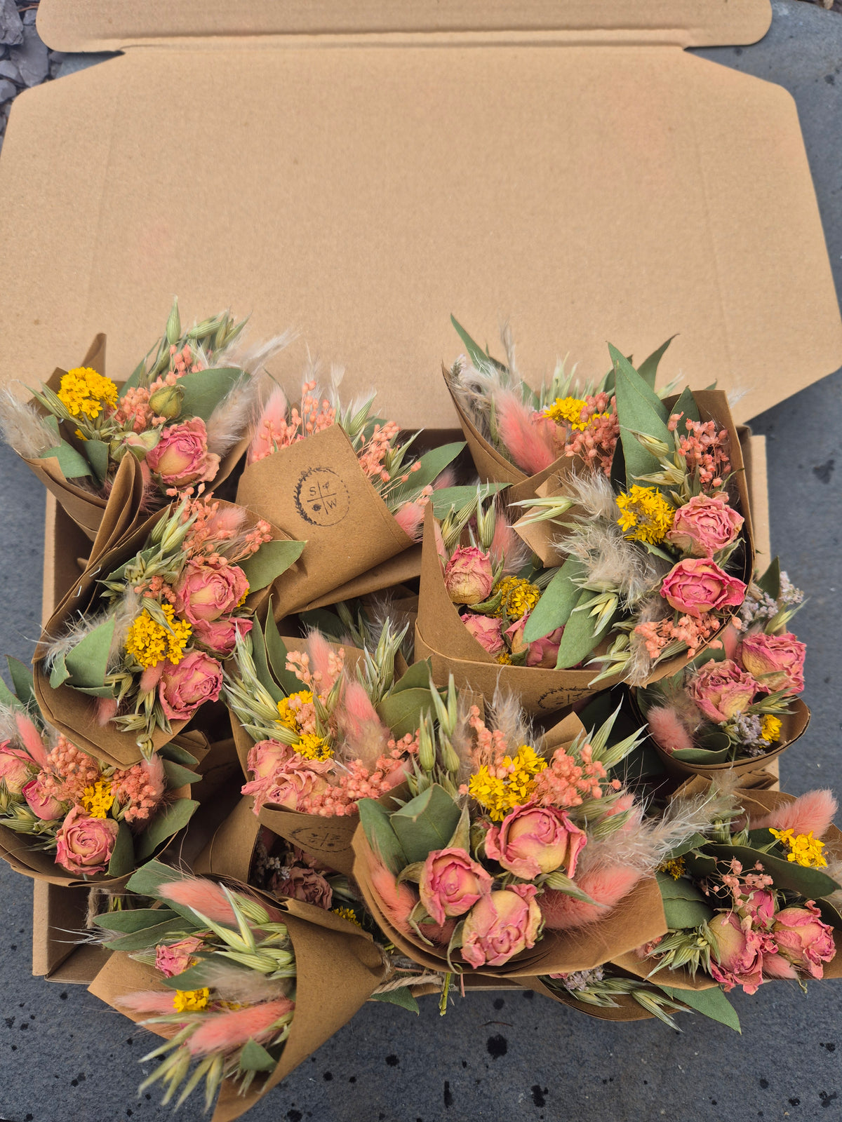 A top-down view of several small dried flower bouquets wrapped in brown kraft paper, arranged inside an open cardboard box on a gray surface.