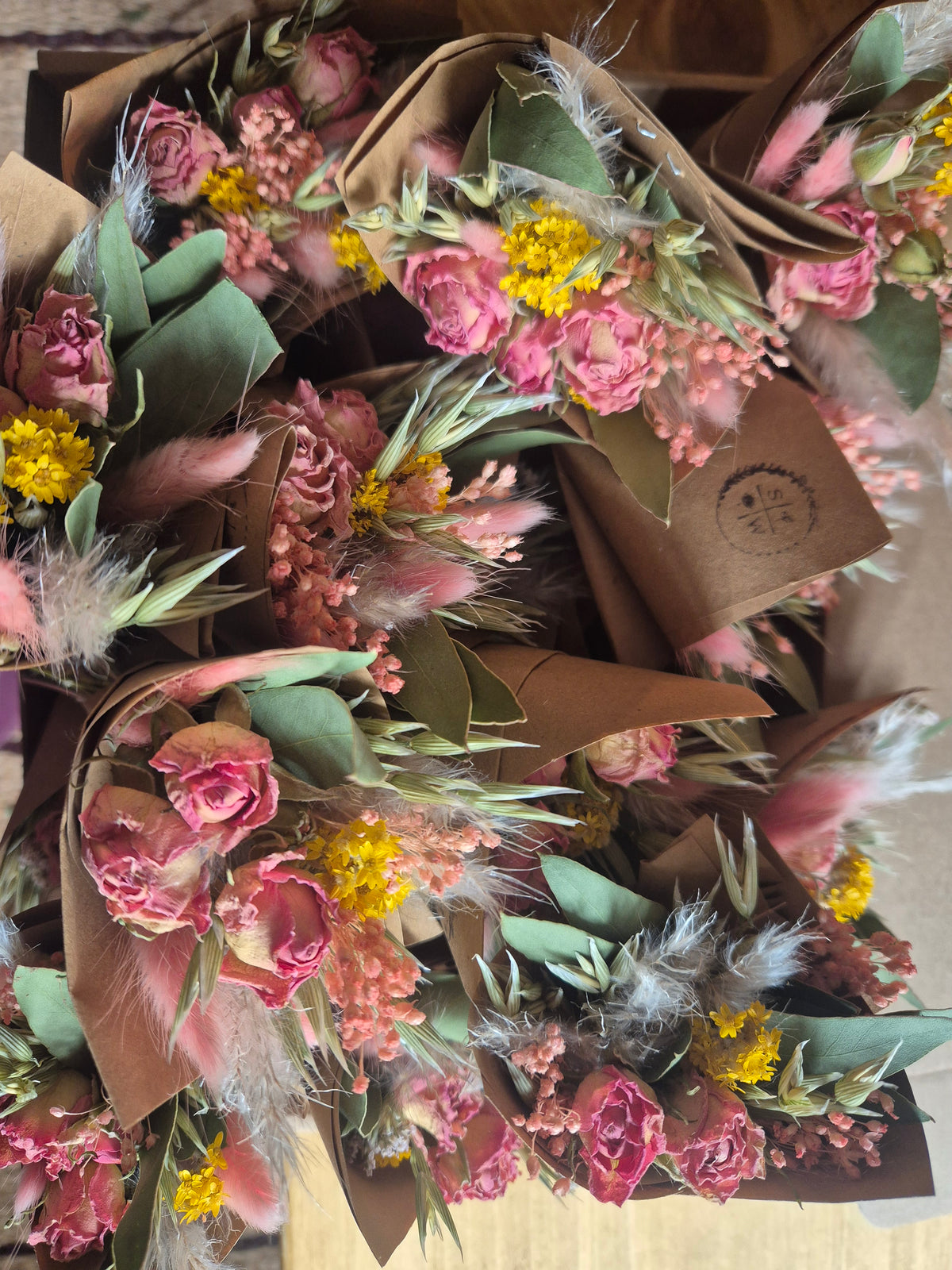 A close-up overhead view of several dried flower bouquets wrapped in brown kraft paper, featuring pink dried roses, yellow dried flowers, green leaves, and fluffy white accents.