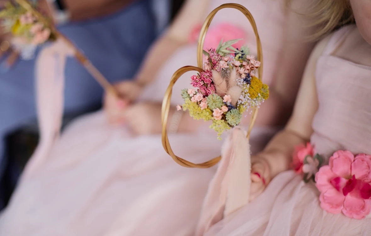 A close-up lifestyle shot of a child in a light pink dress holding a wicker heart-shaped hoop ornament decorated with a small dried flower arrangement. 