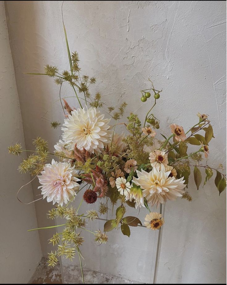 A fresh floral arrangement featuring large cream and white dahlias, delicate pale pink flowers, and green-brown foliage, displayed against a textured off-white wall.