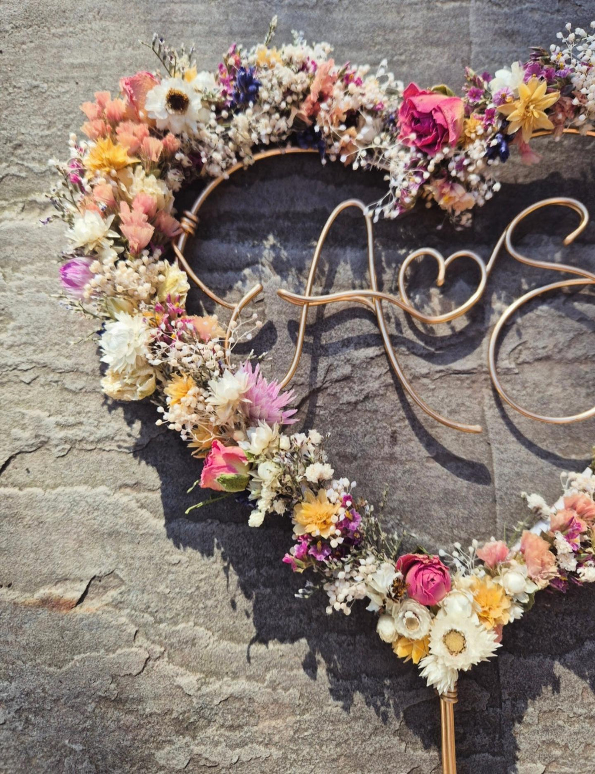 A personalised dried flower cake topper featuring pink, yellow and white hues in the shape a heart.