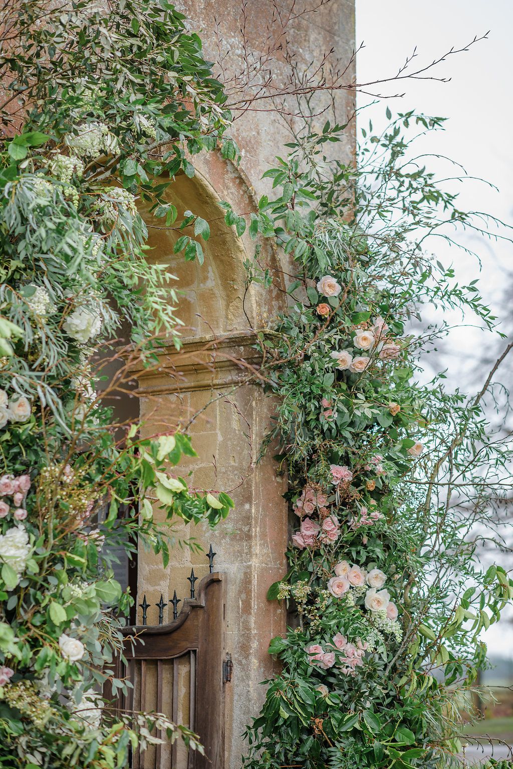 A rustic stone wall with an arched doorway framed by abundant green foliage and soft pink and white flowers.