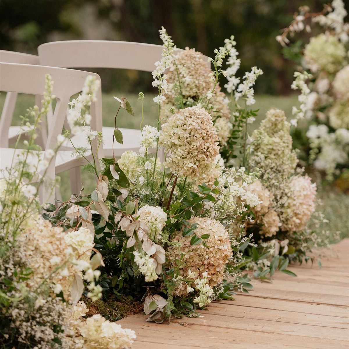 A rustic wedding aisle decorated with lush arrangements of cream and beige hydrangeas, green foliage, and white chairs on a wooden deck outdoors.