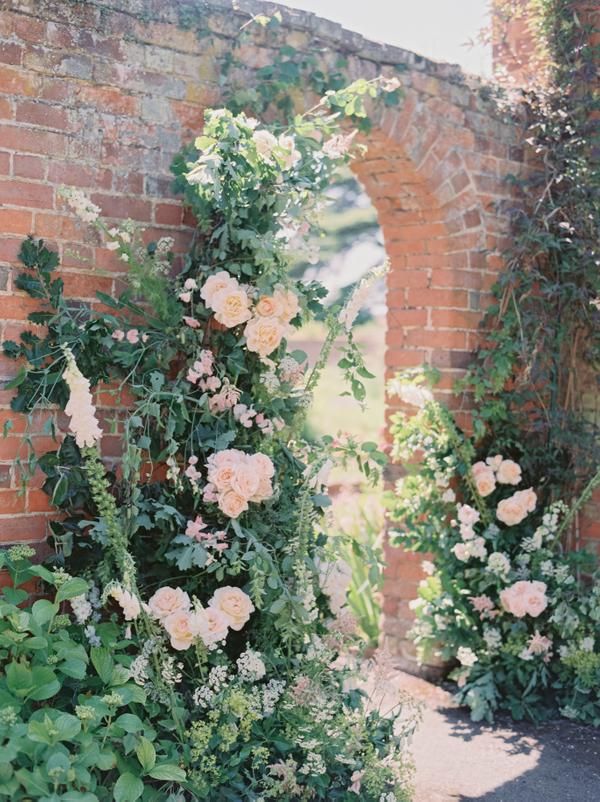 A lush floral archway with pale pink roses and green foliage growing along a rustic red brick archway in a sunlit outdoor garden setting.