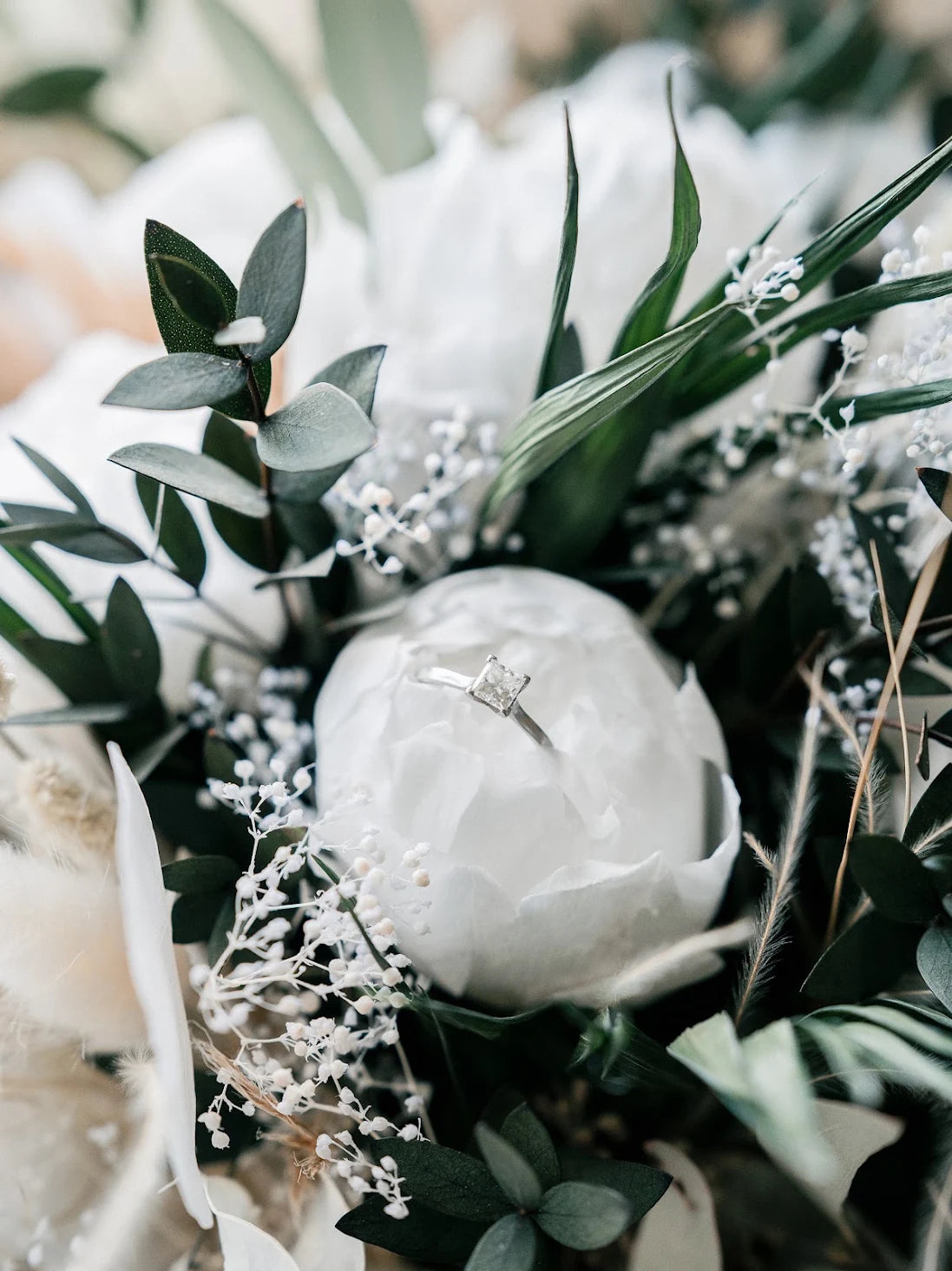 A close-up of a white peony bouquet with green foliage and delicate white baby's breath, featuring a silver engagement ring resting on the center white flower.