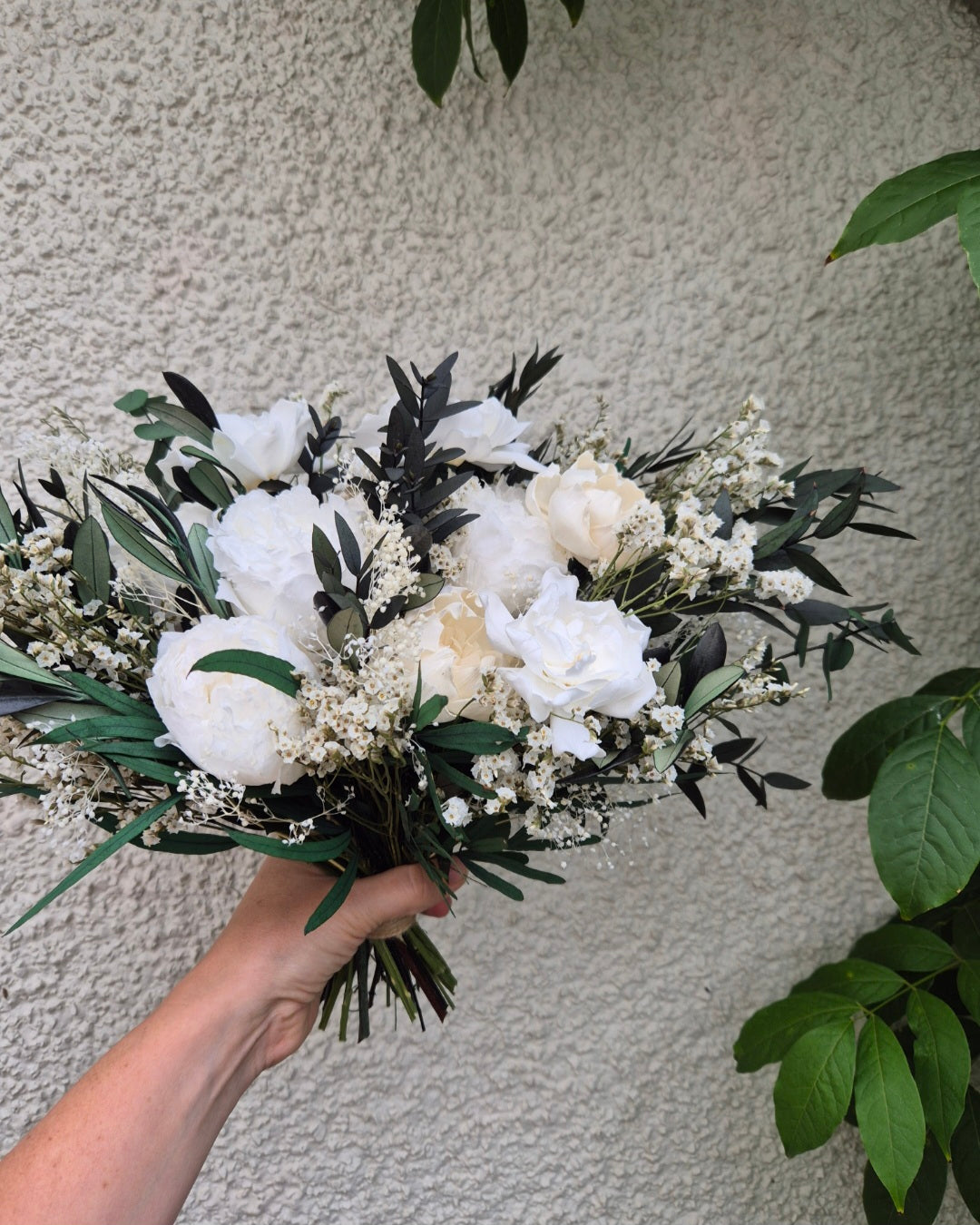 A hand holding a rustic dried flower bouquet featuring white peony roses, cream sola roses, gypsophila, and dark green eucalyptus.