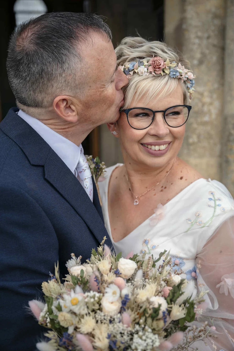 A smiling bride in a white off-the-shoulder wedding dress and floral crown kisses from her groom on the cheek. She is wearing black-rimmed glasses and holding a dried flower bouquet.