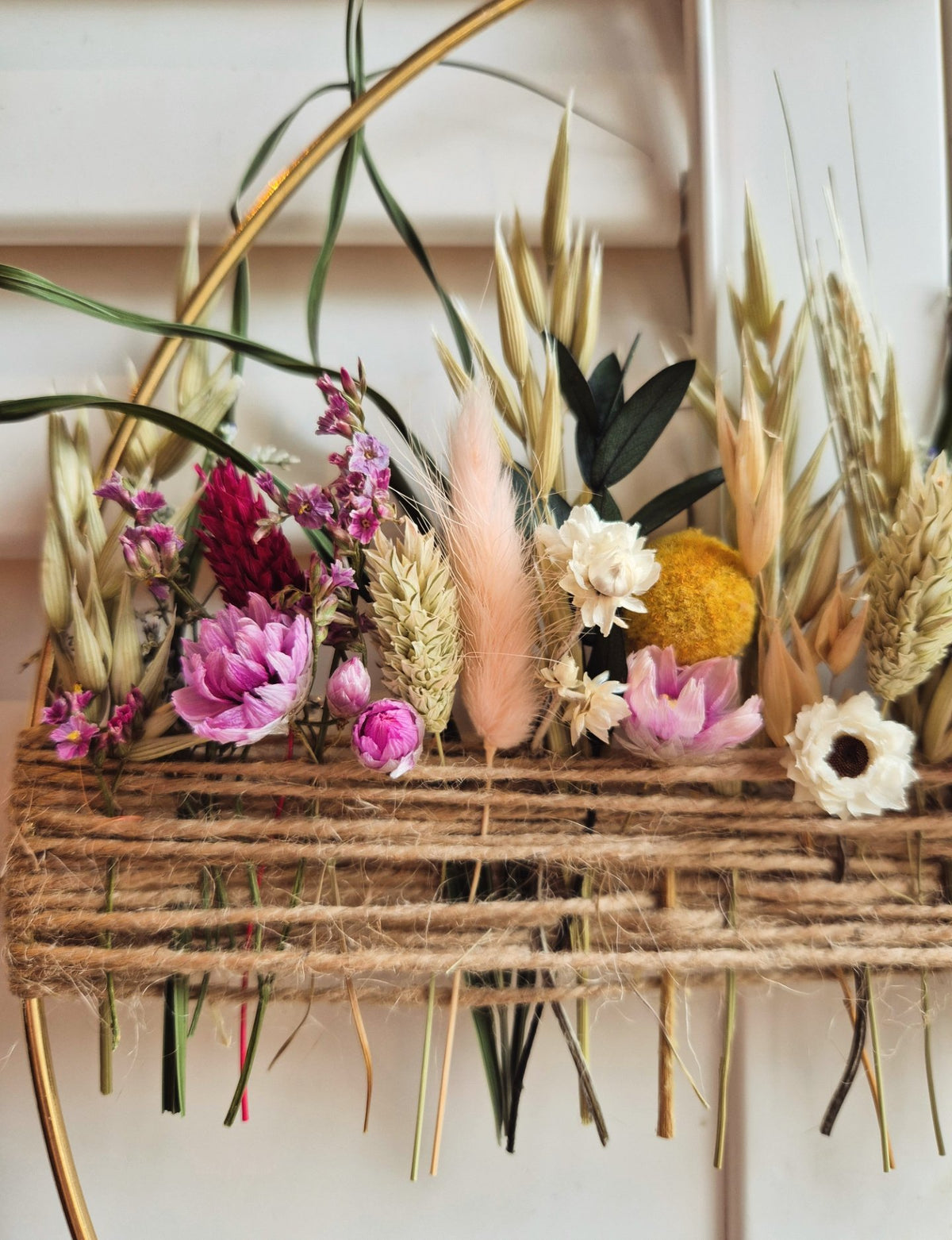 A rustic dried flower and grass arrangement in a woven twine arrangement, featuring pink and white flowers, with wheat, barley and phalaris displayed against a light paneled background.