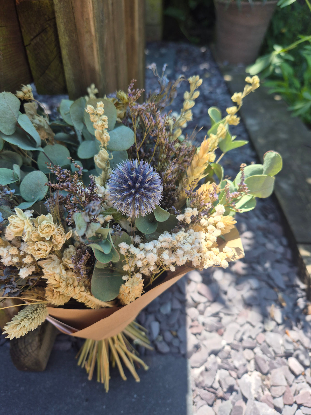 A dried flower bouquet featuring pale cream larkspur, a blue echinop, sage green eucalyptus, and wheat stems, wrapped in brown paper and photographed outdoors on a dark surface with gravel in the background.