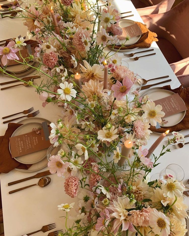 A beautifully styled wedding table centerpiece featuring a lush floral arrangement of pink, white, and yellow wildflowers with lit candles, set on a white tablecloth with brown plates, brown napkins, and gold-toned cutlery.
