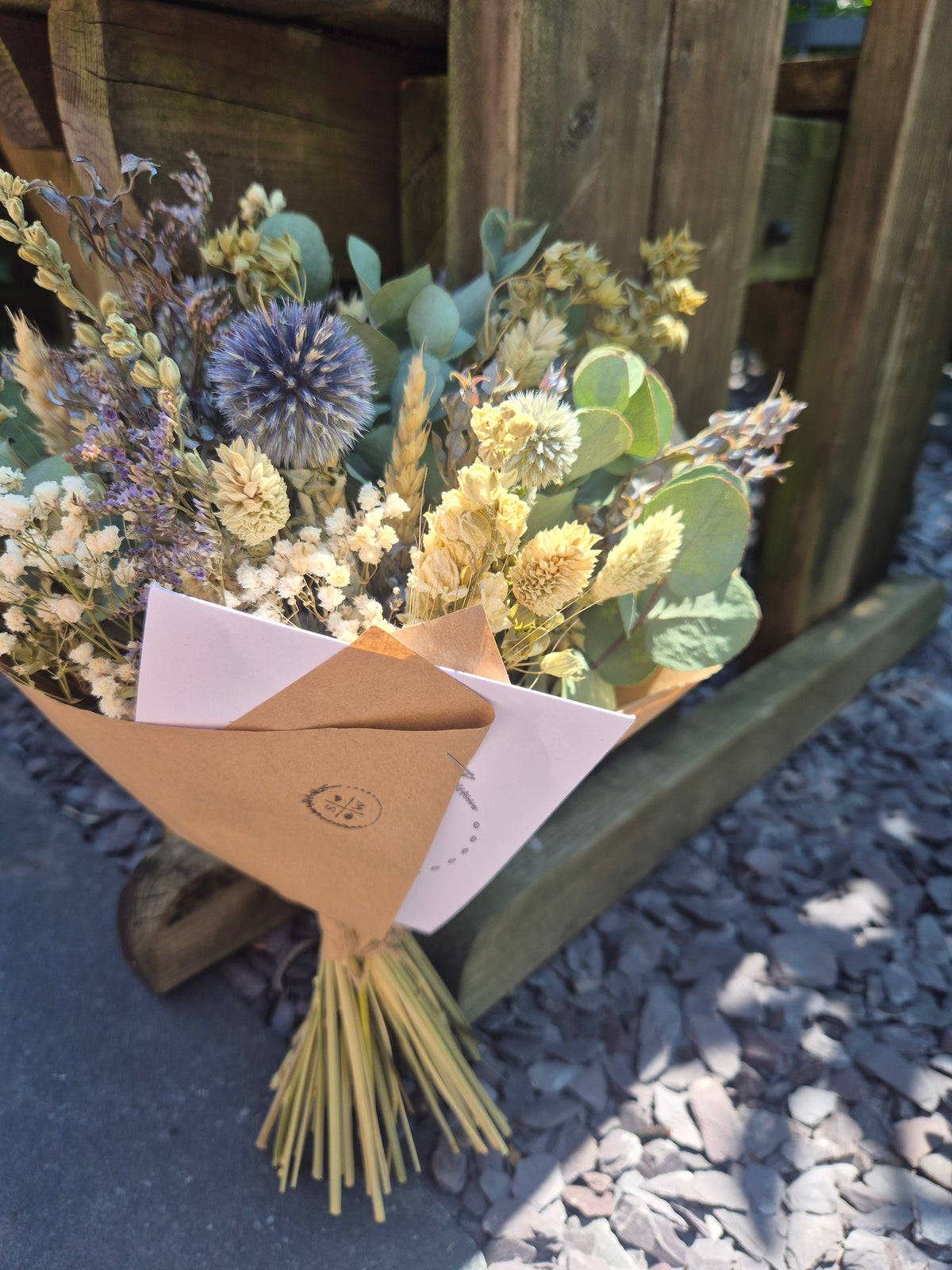 A dried flower bouquet with beige, cream, and lavender elements, wrapped in brown kraft paper and a white envelope, placed on a wooden bench outdoors.
