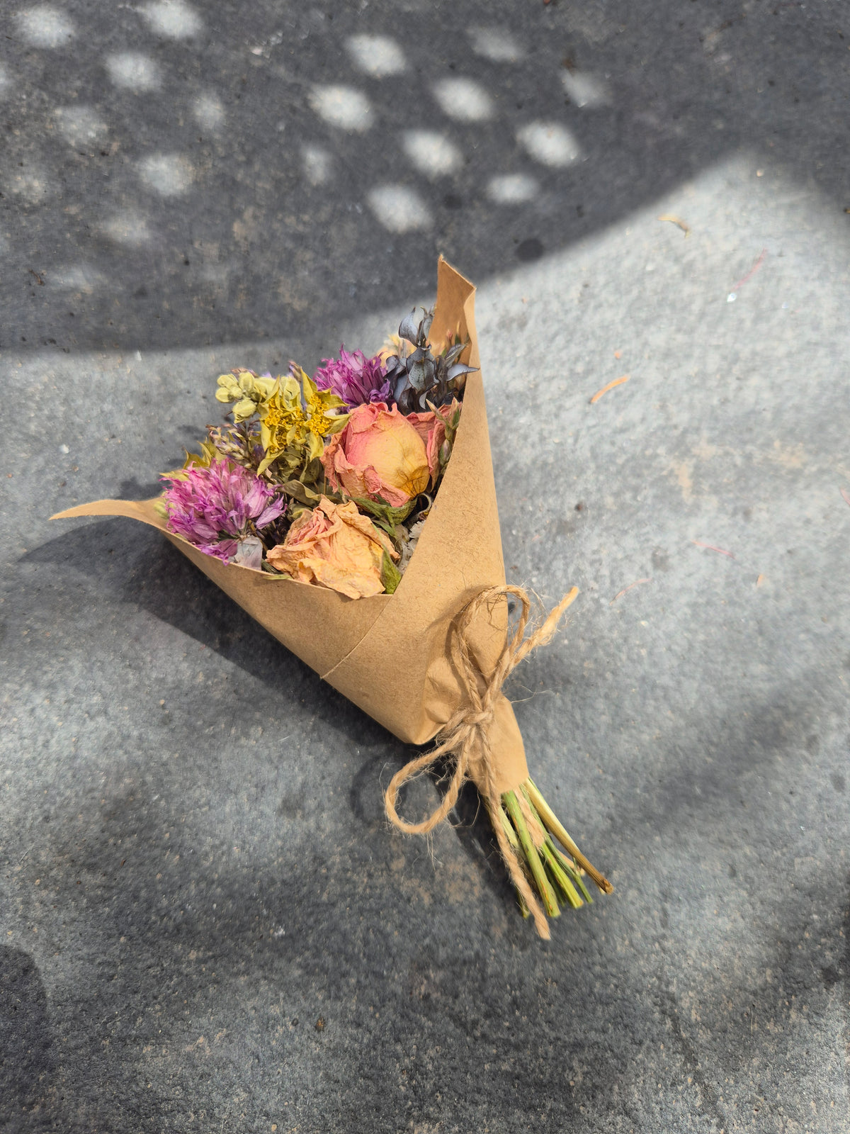 A small bouquet of dried flowers wrapped in brown kraft paper and tied with twine, placed on a gray concrete surface with dappled sunlight and shadows.
